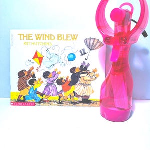 May include: A children's book titled "The Wind Blew" by Pat Hutchins, featuring an illustration of children reaching for items blown by the wind. A pink handheld fan is positioned to the right of the book.