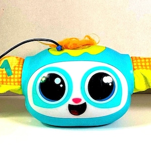 May include: A blue and white baby toy with large black eyes and a smiling face. The toy has orange and yellow wings with numbers and patterns. A black cord extends from the top.
