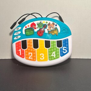 May include: A colorful plastic musical toy with a white base and a teal top section. It features five piano keys labeled with the numbers 1 through 5 in red, orange, yellow, green, and blue. Three instrument icons and cartoon animal characters are visible.
