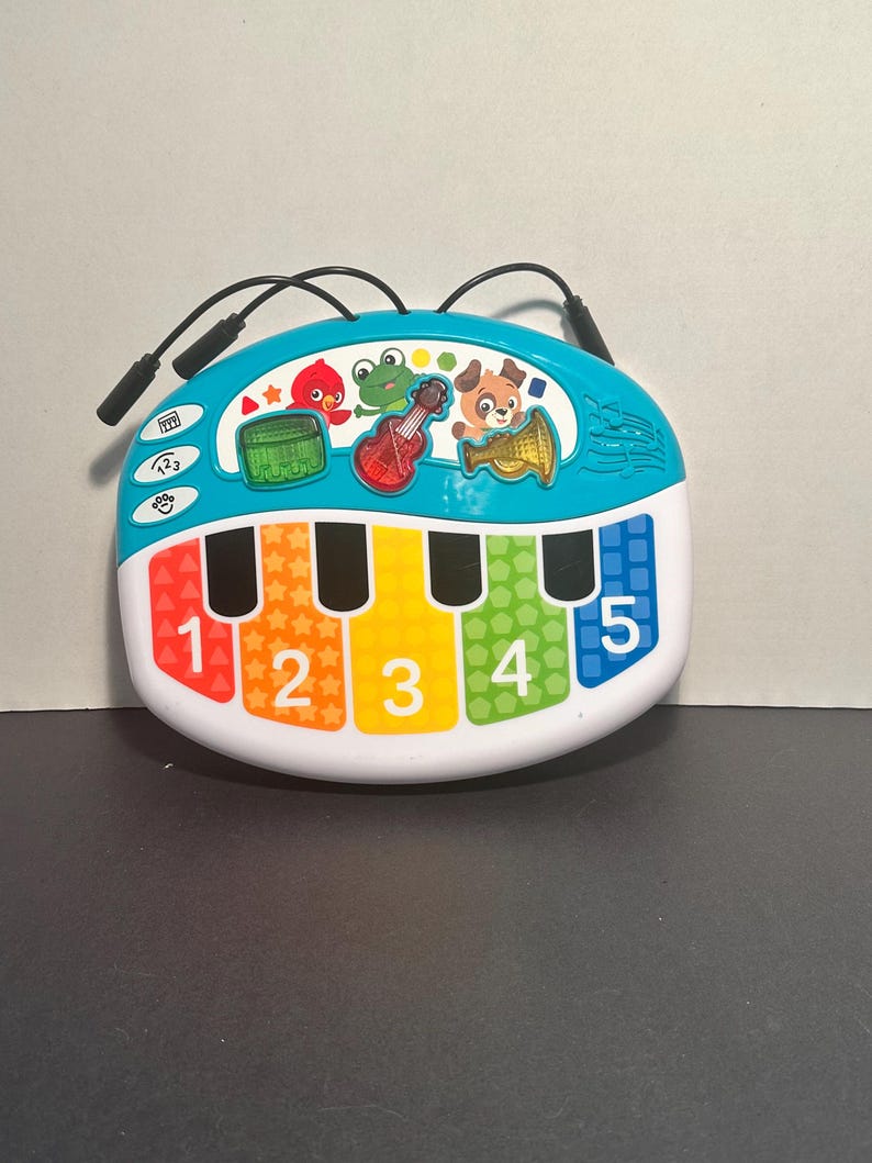 May include: A colorful plastic musical toy with a white base and a teal top section. It features five piano keys labeled with the numbers 1 through 5 in red, orange, yellow, green, and blue. Three instrument icons and cartoon animal characters are visible.
