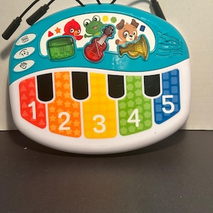 May include: A teal and white musical toy keyboard featuring five colorful keys numbered one through five. The top section displays a red bird, a green frog, and a brown dog, along with a green drum, a red guitar, and a gold trumpet icon.