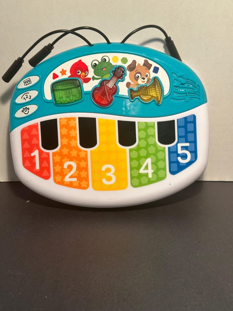 May include: A teal and white musical toy keyboard featuring five colorful keys numbered one through five. The top section displays a red bird, a green frog, and a brown dog, along with a green drum, a red guitar, and a gold trumpet icon.