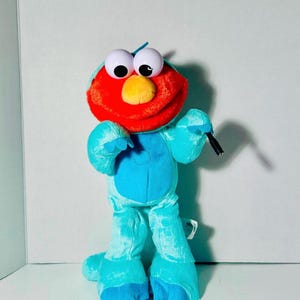 May include: A plush toy of a Sesame Street character, featuring a red head, large white eyes, and a yellow nose. The body is turquoise, with matching feet and hands. The toy is holding a black object.