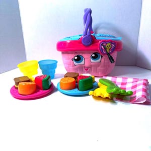 May include: A pink toy picnic basket with a purple handle and a smiling face. The basket is surrounded by toy food items, including miniature cakes, watermelon slices, and orange slices on small plates, along with yellow and blue cups and a pink and white checkered napkin.