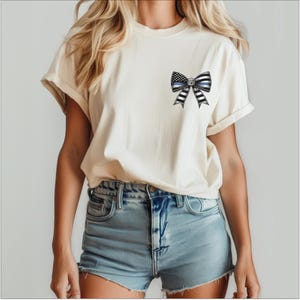Puede incluir: Camiseta color crema con un dise&ntilde;o de lazo que incorpora una l&iacute;nea azul fina y elementos de la bandera estadounidense. El lazo es de rayas blancas y negras. La camiseta se combina con pantalones cortos vaqueros.