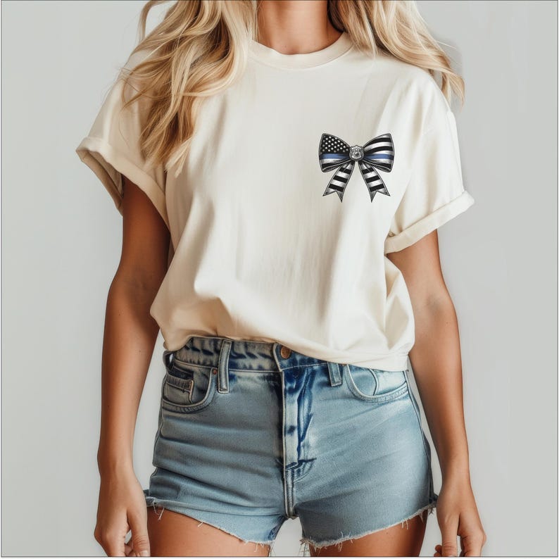 Puede incluir: Camiseta color crema con un dise&ntilde;o de lazo que incorpora una l&iacute;nea azul fina y elementos de la bandera estadounidense. El lazo es de rayas blancas y negras. La camiseta se combina con pantalones cortos vaqueros.