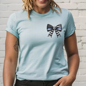 Puede incluir: Camiseta azul claro con un dise&ntilde;o de lazo con la bandera estadounidense y una l&iacute;nea azul fina. El lazo tiene rayas blancas y negras. La camiseta tiene mangas cortas y cuello redondo.