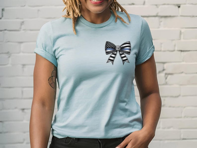 Puede incluir: Camiseta azul claro con un dise&ntilde;o de lazo con la bandera estadounidense y una l&iacute;nea azul fina. El lazo tiene rayas blancas y negras. La camiseta tiene mangas cortas y cuello redondo.