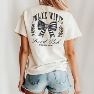 Puede incluir: Camiseta color crema con el texto "POLICE WIVES Social Club" y "BACK THE BADGE". El dise&ntilde;o presenta un lazo con una l&iacute;nea azul y un patr&oacute;n de bandera estadounidense. La camiseta tiene mangas cortas y un corte holgado.