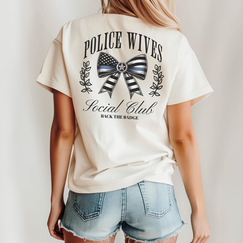 Puede incluir: Camiseta color crema con el texto "POLICE WIVES Social Club" y "BACK THE BADGE". El dise&ntilde;o presenta un lazo con una l&iacute;nea azul y un patr&oacute;n de bandera estadounidense. La camiseta tiene mangas cortas y un corte holgado.