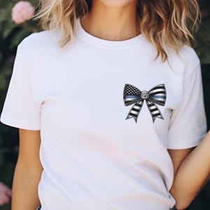 Puede incluir: Camiseta blanca con un dise&ntilde;o de lazo que incorpora una l&iacute;nea azul fina y elementos de la bandera estadounidense. El lazo tiene rayas blancas y negras y una insignia en el centro. La camiseta es de manga corta.