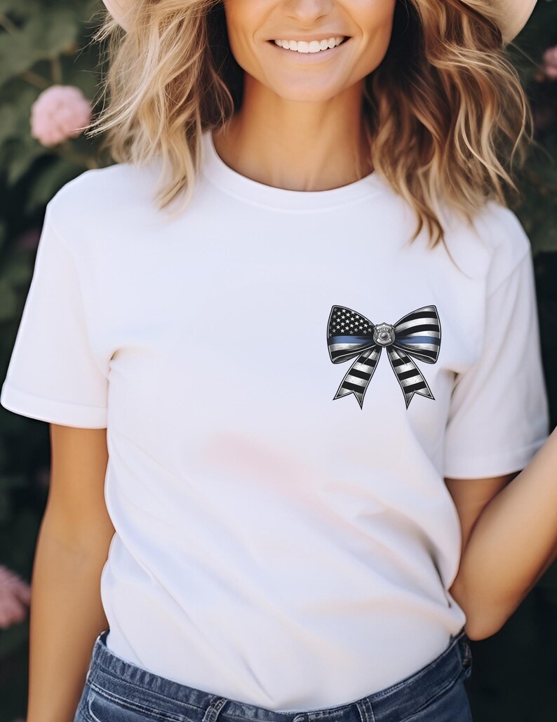 Puede incluir: Camiseta blanca con un dise&ntilde;o de lazo que incorpora una l&iacute;nea azul fina y elementos de la bandera estadounidense. El lazo tiene rayas blancas y negras y una insignia en el centro. La camiseta es de manga corta.