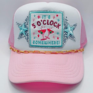 May include: Pink and white trucker hat with a pink brim and mesh back. The hat features a patch with the text "It's 5 O'Clock Somewhere!" and two cocktail glasses. The patch is surrounded by glittery stars and a gold chain.