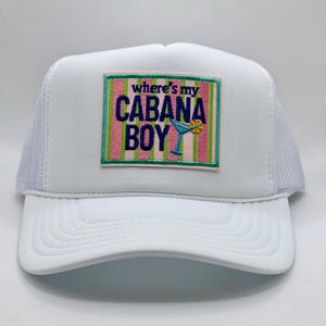 May include: A white trucker hat featuring a rectangular embroidered patch on the front. The patch has vertical pink and green stripes with the text "where's my CABANA BOY" in dark blue, accompanied by a small blue cocktail glass with a yellow slice.