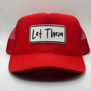 May include: A red trucker hat with a mesh back and a curved brim. The hat features a white rectangular patch with the words "Let Them" in black script.