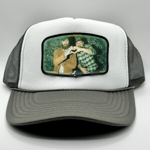 May include: A white and gray trucker hat with a black patch featuring a photo of two people making a heart shape with their hands.
