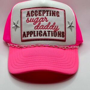 May include: A pink and white trucker hat with a silver chain. The hat has a white patch with pink glitter text that reads "ACCEPTING sugar daddy APPLICATIONS".