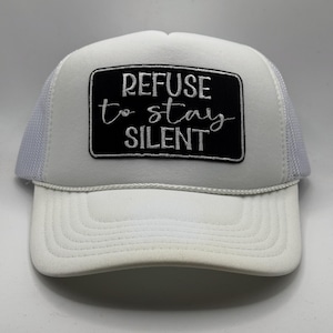 May include: White trucker hat with a mesh back and a curved brim. The front features a black rectangular patch embroidered with the words "REFUSE to STAY SILENT" in white script and block letters. The hat has a white foam front.