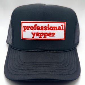 May include: Black trucker hat with a mesh back and a curved brim. The hat features a rectangular patch with the words "professional yapper" in red lettering on a white background, framed by a red border.