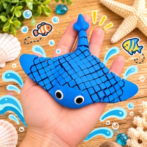 May include: A blue articulated stingray toy with a segmented body and tail, featuring two large black and white eyes. The surface of the stingray is composed of a grid of small square sections, allowing for flexible movement of the toy.