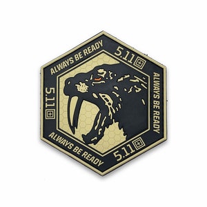 Może przedstawiać: Heksagonalna naszywka w kolorze czarnym i brązowym z grafiką wściekłego szablastozębnego kota. Naszywka zawiera napis "5.11" i "ALWAYS BE READY" powtórzony trzy razy wokół obwodu.