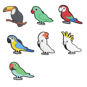Puede incluir: Una colección de siete amuletos coloridos con forma de pájaro. Los amuletos incluyen un tucán, loros verdes, un loro rojo, un loro azul y amarillo y cacatúas blancas con crestas naranjas y amarillas. Cada amuleto tiene una base negra.