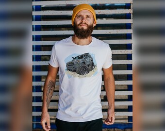 Op de afbeelding: Een wit T-shirt met een cartoonillustratie van een grijze terreinwagen met zwarte banden. De auto rijdt over een onverhard pad.