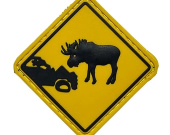 May include: Yellow diamond-shaped warning sign with black border and stitching. The sign depicts a black silhouette of a moose and a car. The sign reads "Moose Crossing".