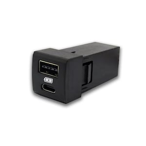 Könnte beinhalten: Schwarzer Kunststoff-Autoladeadapter mit zwei Anschlüssen: ein USB-A-Anschluss und ein USB-C-Anschluss. Der USB-A-Anschluss hat ein Blitzsymbol und der USB-C-Anschluss ein Batteriesymbol.