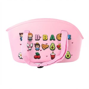 Puede incluir: Un bolso tote rosa con una parte superior curvada y un asa rosa. La bolsa est&aacute; decorada con im&aacute;genes coloridas de estilo caricatura y la palabra "BBAGE" en letras individuales. La bolsa tiene un cierre negro.