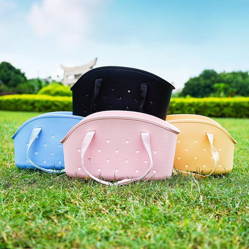 Puede incluir: Cuatro bolsas de mano coloridas en negro, azul, rosa y amarillo est&aacute;n dispuestas sobre hierba verde. Cada bolsa tiene una parte superior curva, dos asas y recortes en forma de coraz&oacute;n. Las bolsas est&aacute;n hechas de un material suave y flexible.