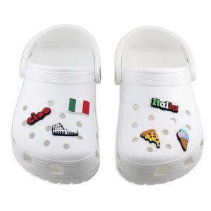 May include: White clogs decorated with Italian-themed charms. One clog features a flag, a Roman Colosseum charm, and the word "ciao." The other clog has a pizza slice, an ice cream cone, and the word "Italia."