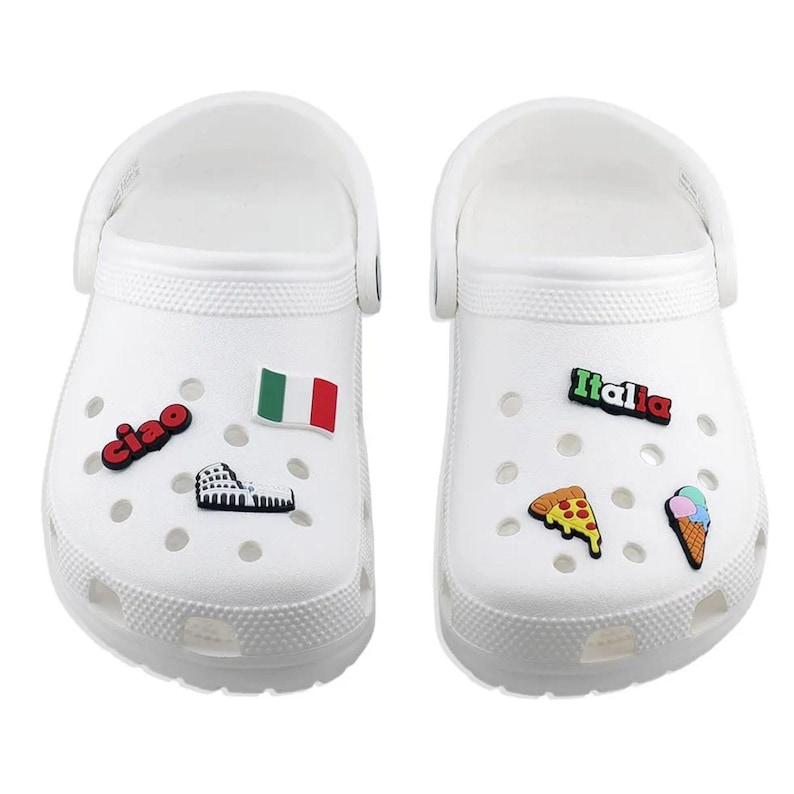 May include: White clogs decorated with Italian-themed charms. One clog features a flag, a Roman Colosseum charm, and the word "ciao." The other clog has a pizza slice, an ice cream cone, and the word "Italia."