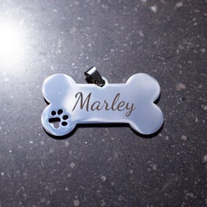 以下が含まれることがあります： 骨の形をした、光沢のある銀色の犬用ネームタグ。「Marley」という名前が筆記体で刻まれています。左側には足跡がエッチングされています。小さな金属製のループが付いています。