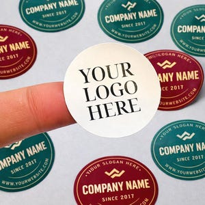 Peut inclure: Gros plan sur plusieurs autocollants ronds. L'un d'eux, tenu par un doigt, porte le texte "YOUR LOGO HERE" en noir. D'autres autocollants affichent "COMPANY NAME" et "YOUR SLOGAN HERE" en blanc et or.