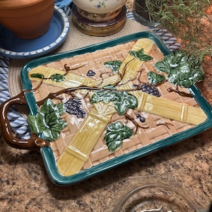 May include: Rectangular ceramic serving tray with a woven basket design, green trim, and a brown handle. The tray features raised relief details of grape vines, leaves, and bunches of grapes in shades of green, yellow, and purple.