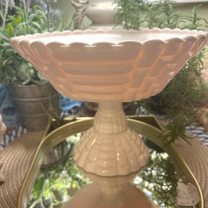 May include: A light pink ceramic cake stand with a scalloped edge and a textured basketweave design. The stand has a pedestal base and is displayed on a gold-rimmed mirror. The stand is surrounded by greenery and other decorative items.