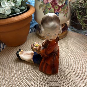May include: A ceramic figurine of a child sitting with a bouquet of flowers in their hand. The figurine is painted in shades of brown, red, blue, and white.