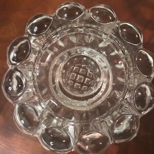 May include: Clear glass ashtray with a circular design. The ashtray features a series of rounded, bubble-like protrusions around the rim. The center has a textured, grid-like pattern. The glass is transparent, allowing light to pass through.