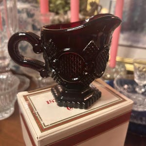 May include: A ruby red glass creamer with a decorative handle and ornate embossed detailing. The creamer sits on a white box with red stripes and the number "187" visible. The creamer is likely vintage or antique.
