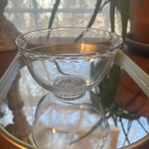 May include: A clear glass bowl with a decorative etched rim, resting on a mirrored surface. The bowl's design features a delicate pattern around the top edge. The bowl is likely used for serving small portions of food or as a decorative item.