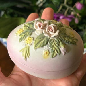 May include: A pale pink, oval-shaped trinket box. The lid is adorned with a detailed floral arrangement featuring two pink roses, green leaves, and small yellow flowers. The box is held in a hand.