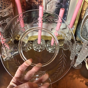 May include: A clear glass plate with etched leaf designs. The plate is held up in front of pink candles and decorative candle holders. The plate has a circular design with a detailed leaf pattern around the edge.