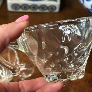 May include: Clear glass teacup with a textured, swirled design. The cup has a handle and a scalloped base. The image shows a close-up of the cup, highlighting its details. The teacup is part of a tea set.