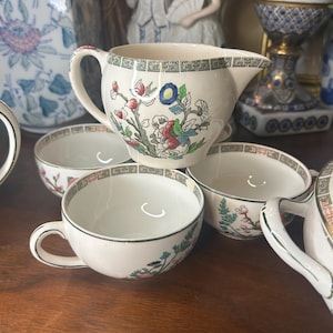 May include: Cream-colored ceramic tea set featuring floral designs in red, blue, and green. The set includes a creamer and several teacups. The rims and handles are accented with green trim. The tea set is placed on a wooden surface.
