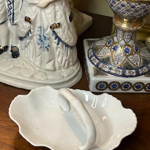 May include: A white porcelain serving dish with a scalloped edge and a curved handle. The dish is oval-shaped and sits on a wooden surface. Other porcelain figurines and decorative items are in the background.