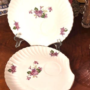 May include: Two white porcelain plates with a scalloped edge and a floral violet design. Each plate features a central cluster of purple violets with green leaves and smaller violet accents. The plates have a gold trim.