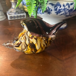 May include: A decorative glass seashell-shaped bowl with a dark purple and yellow swirl pattern. The bowl has a clear, pointed spout and a matching lid. The bowl is sitting on a wooden surface.