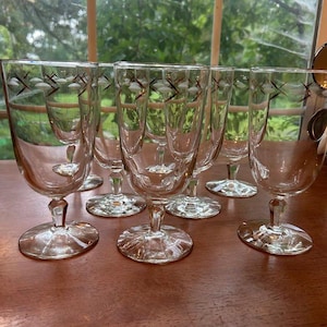 May include: A set of clear glass goblets with a decorative pattern of white and brown cross-like shapes. The goblets have a stemmed base and are arranged on a wooden surface, suggesting a vintage or antique aesthetic.