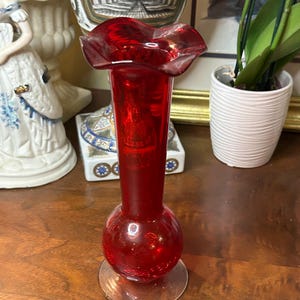 May include: A vibrant red glass vase with a ruffled top and a bulbous base. The vase has a clear, circular base and stands on a wooden surface. Decorative items are in the background.